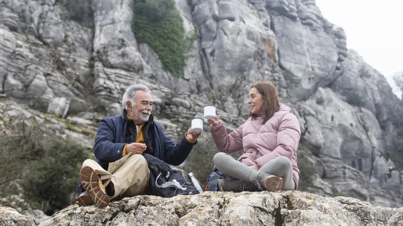 Dos senderistas mayores sentados en una roca en la montaña brindan con sus tazas