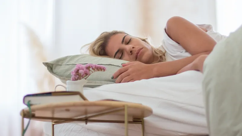 Mujer duerme entre sábanas blancas, con un libro y flores al lado