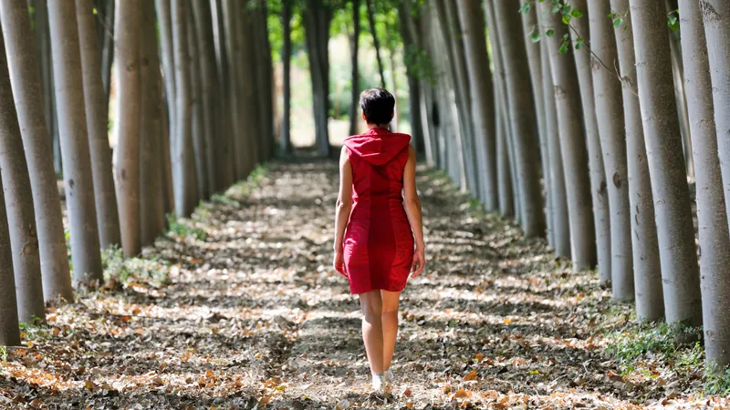 Mujer con vestido rojo camina de espaldas por un camino cubierto de hojas entre dos hileras de árboles altos