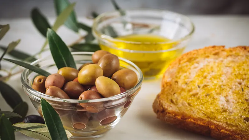 Pan tostado junto a un cuenco de aceitunas y otro de aceite de oliva, con ramas de olivo sobre la mesa